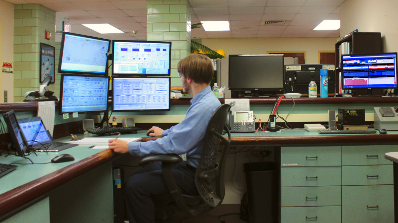 Brunette man sits in front of 6 computer monitors, operating the water treatment plant.