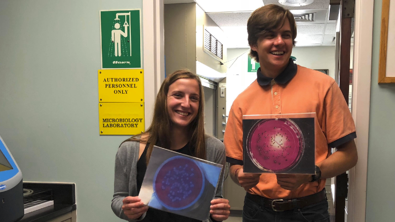 Two college interns hold photos of microbes and smile.