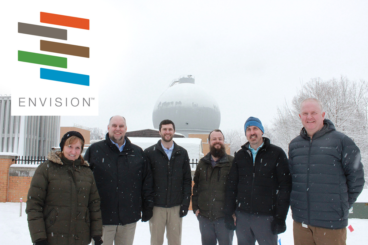 Six adults standing in front of a wastewater plant. The Envision logo is in the upper left corner.