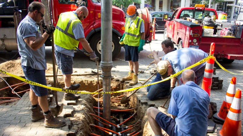 Men in blue work uniforms and high vis vests dig a hold in a sidewalk surrounded by red BPW trucks