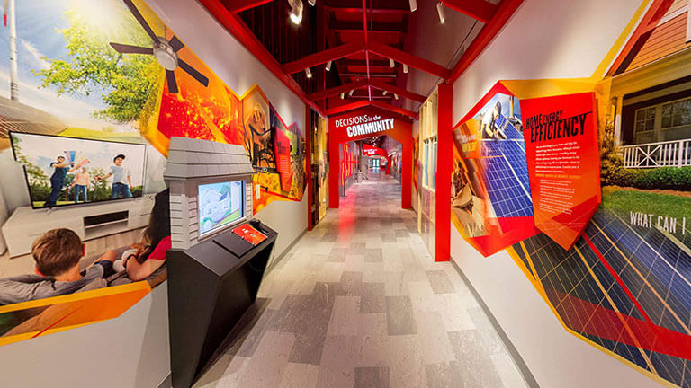 A brightly-lit hallway decorated with warm colored geometric museum displays and a house-shaped kiosk on the left.