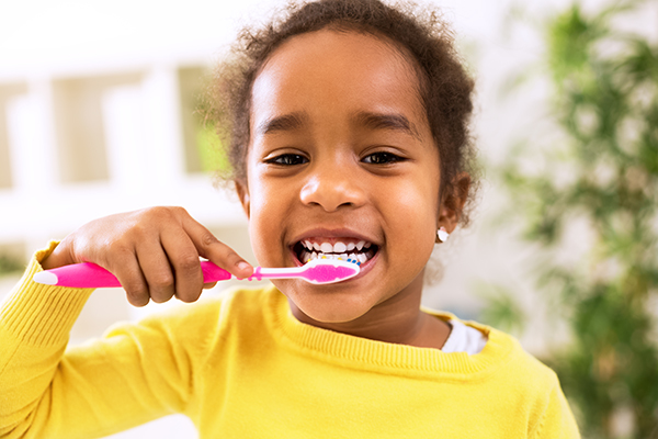 Little girl in a yellow shirt brushes her teeth with a pink toothbrush.