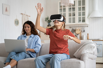 a girl sits on a couch actively playing a VR game. Her mother sits next to her working on her laptop