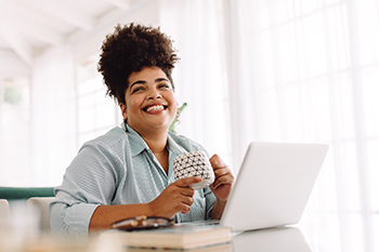 woman smiling sits in front of a laptop holding a cup of coffee