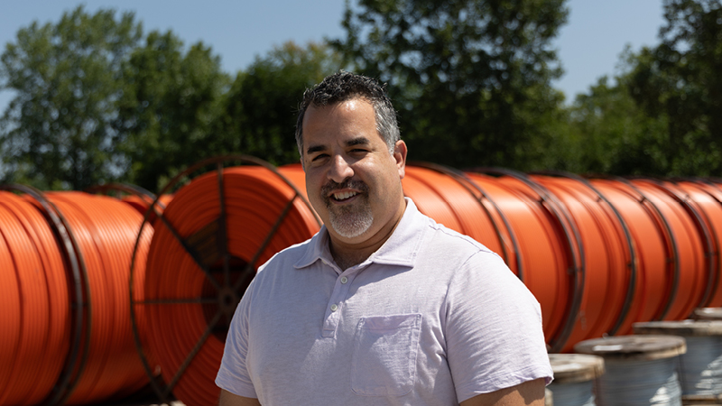 middle age man with dark hair stands in front of orange rolls of fiber conduit