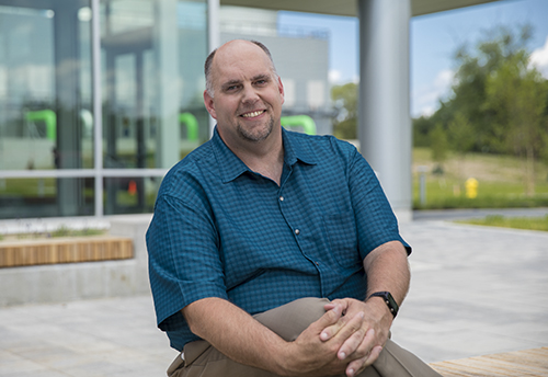 General Manager Dave Koster sits in front of Holland Energy Park wearing a teal button-down shirt with hands over his crossed knees.