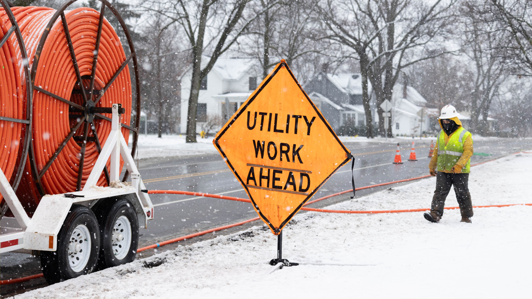 a city street with a sign that says utility work ahead and a utility worker walking towards trailer holding orange spools of fiber conduit